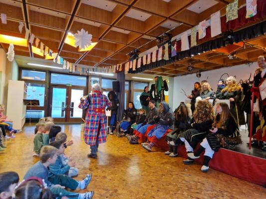 Person in bunter Kleidung steht in einem Saal vor sitzenden Kindern und Erwachsenen, die auf einer Bühne und am Boden sitzen
