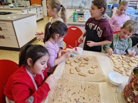 Kinder sitzen und stehen an einem Tisch und verzieren mit Spritzglasur und Streuseln verschiedene Plätzchen auf Backpapier