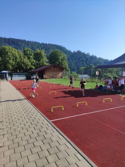 Außenansicht einer Sportanlage mit roter Laufbahn, angrenzendem gepflastertem Bereich und bewaldetem Hintergrund.