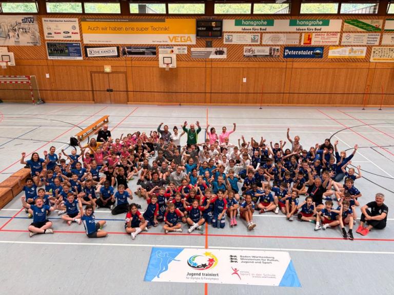Gruppenfoto in einer Sporthalle: Viele Kinder und einige Erwachsene sitzen und stehen gemeinsam auf dem Hallenboden und winken in die Kamera.