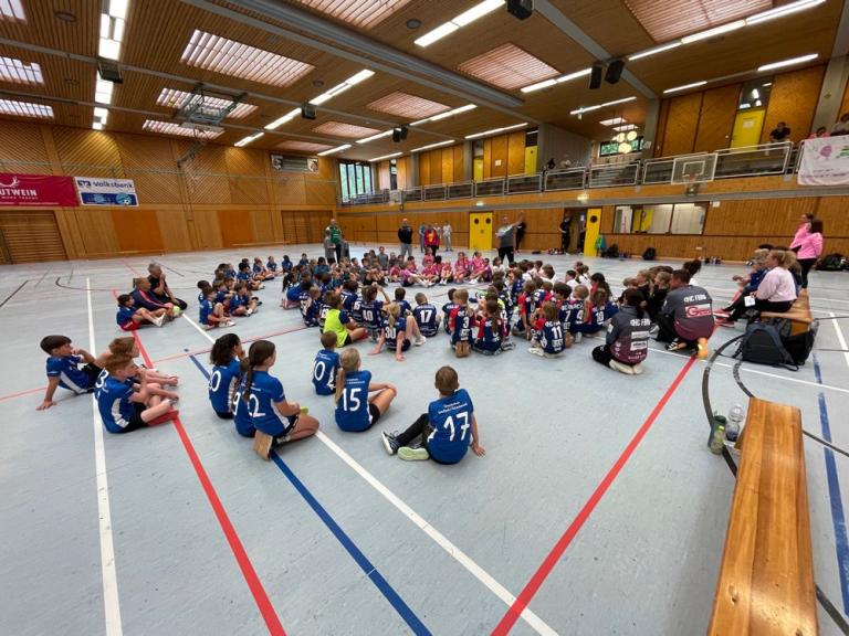 Kinder in Sportkleidung sitzen in einer Turnhalle im Kreis, während einige Erwachsene in der Mitte stehen und sprechen.