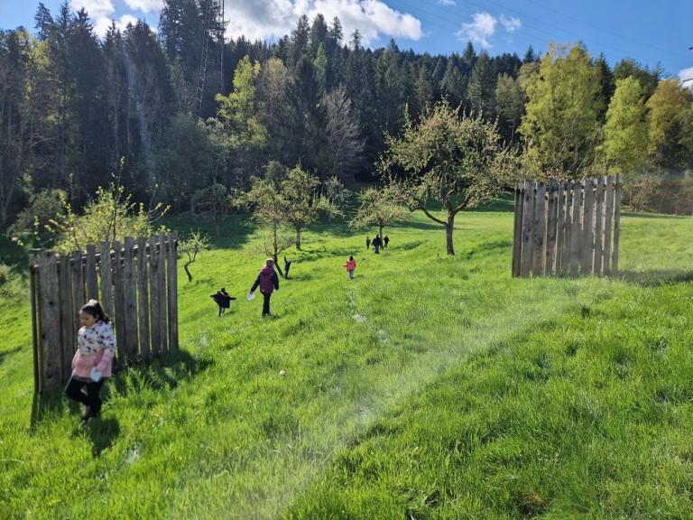 Kinder und eine erwachsene Person laufen über eine sonnige Wiese zwischen blühenden Obstbäumen; im Hintergrund liegt ein Waldrand.