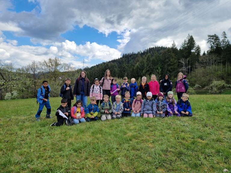 Gruppe von Kindern und zwei erwachsenen Personen posiert auf einer Wiese vor einem bewaldeten Hang unter einem teils bewölkten Himmel.