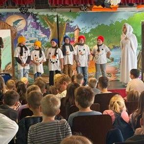 Kinder in Kostümen stehen auf einer Bühne vor einem gemalten Bühnenbild und einem Publikum aus Kindern