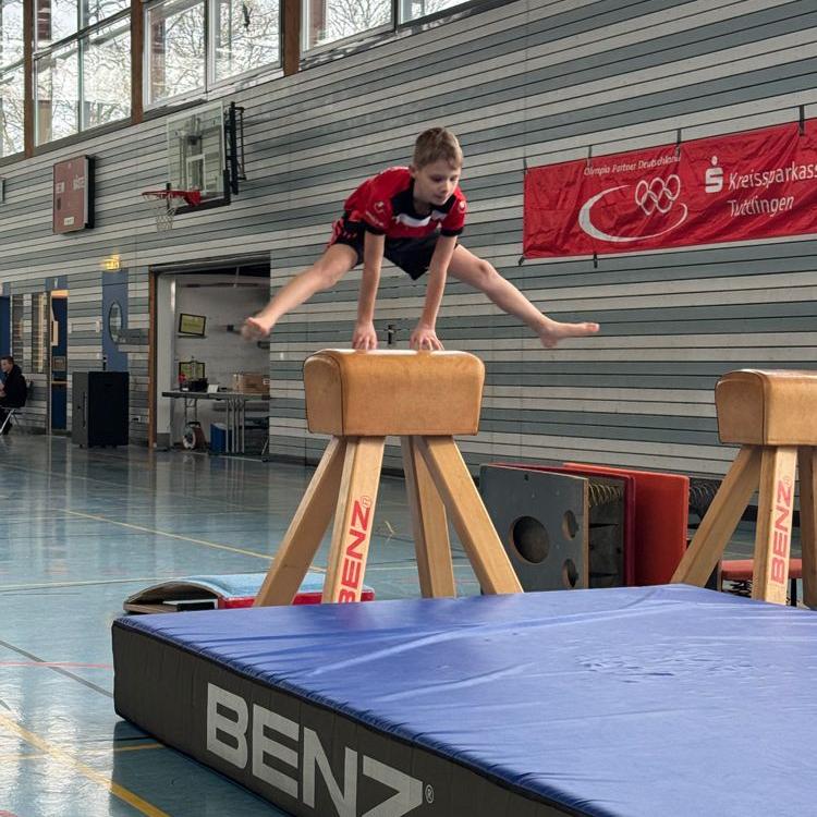 Kind in rotem Trikot macht einen Spreizsprung über einen Turnpferd in einer Sporthalle mit blauer Matte und Banner im Hintergrund